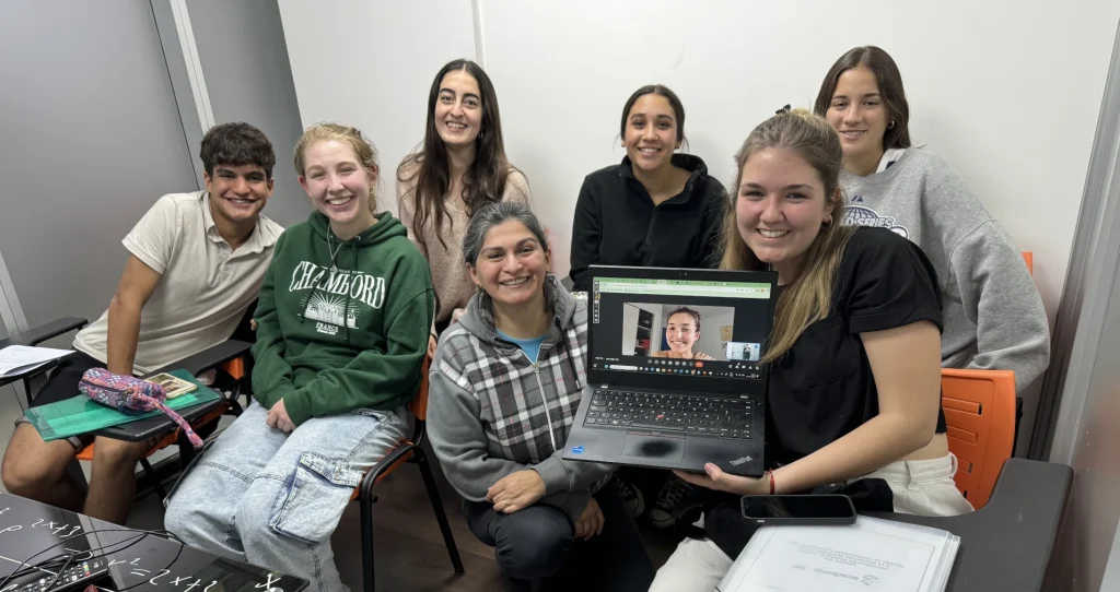 Grupo de estudiantes sonriendo en una sala de clases junto docente Marcela Guerra y sosteniendo una laptop con una videollamada activa.