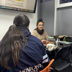 Una estudiante está sentada en una mesa, recibiendo tutoría o apoyo adicional de una profesora. Están frente a un monitor de TV que muestra texto.