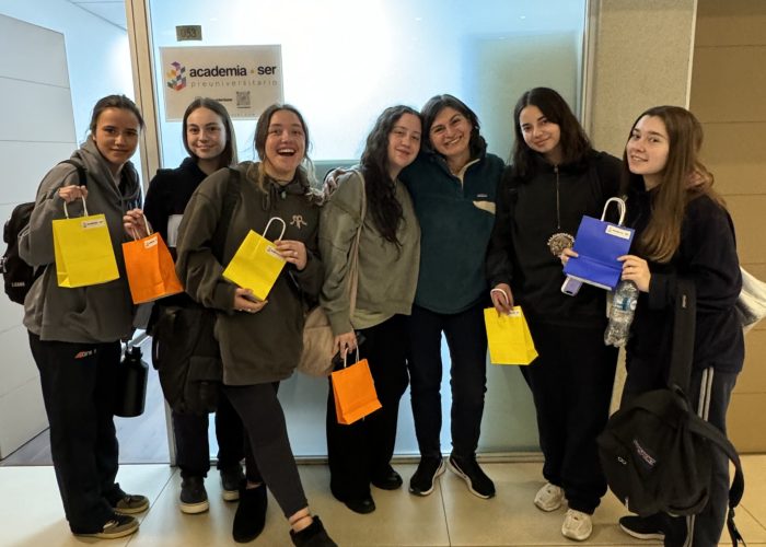 Grupo de seis estudiantes sonrientes junto a docente Marcela Guerra, posando juntos frente a un letrero de Preuniversitario Ser, sosteniendo pequeñas bolsas de regalo de colores amarillo, naranja y azul.