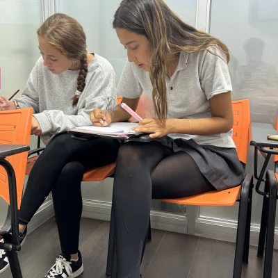 Dos estudiantes sentadas en sillas naranjas dentro de una sala de clases, concentradas escribiendo y tomando apuntes en sus cuadernos.